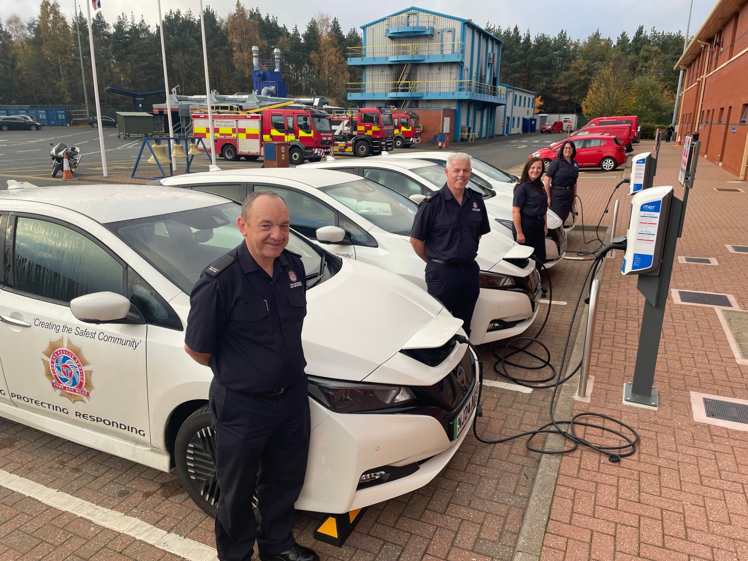 Fire and Rescue Employees standing next to a fleet of electric vehicles at a charging point. Fire Appliances are in the background of the image.