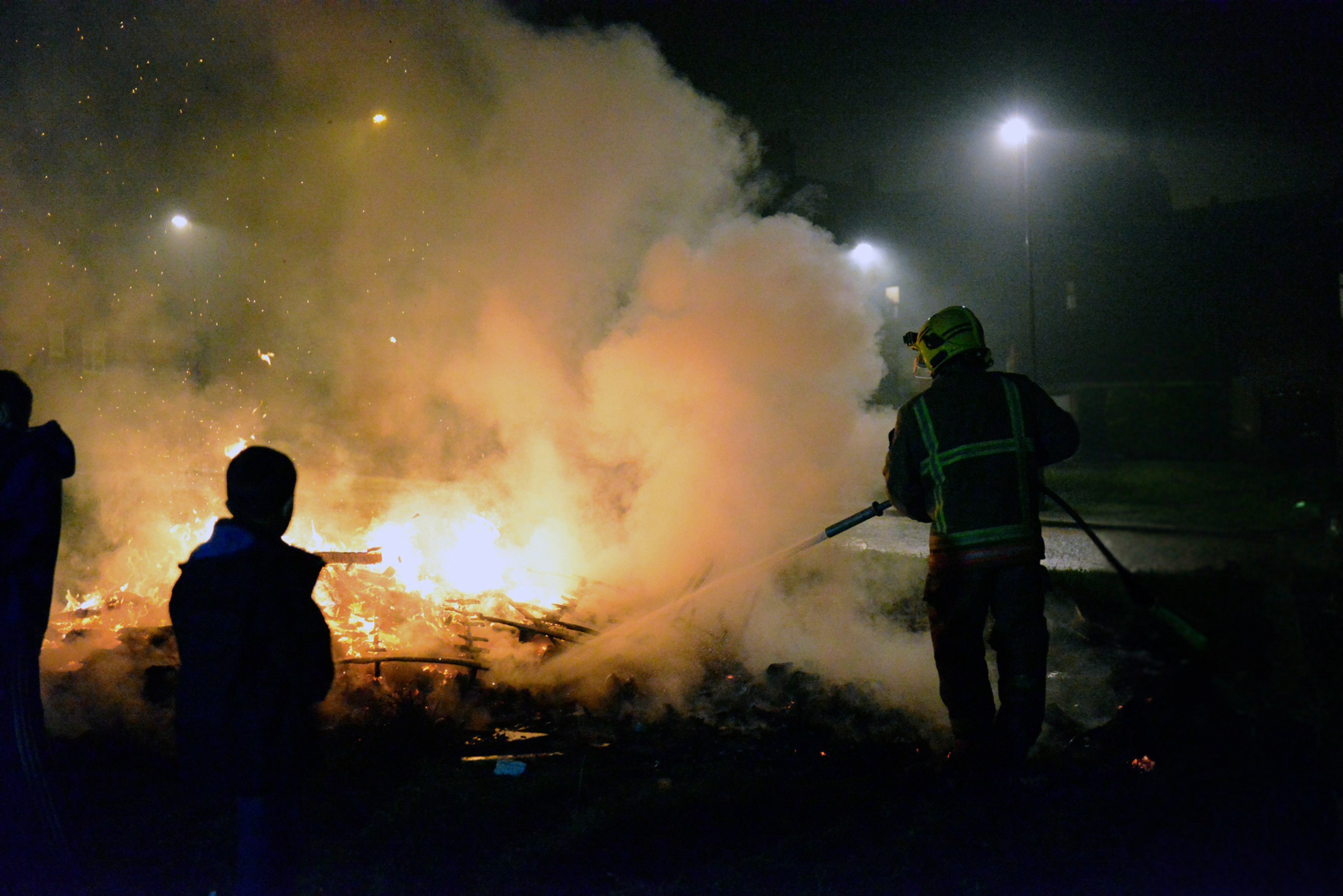 Image of firefighter using hose reel to extinguish fire in residential area. Youths seen in forefront of image.