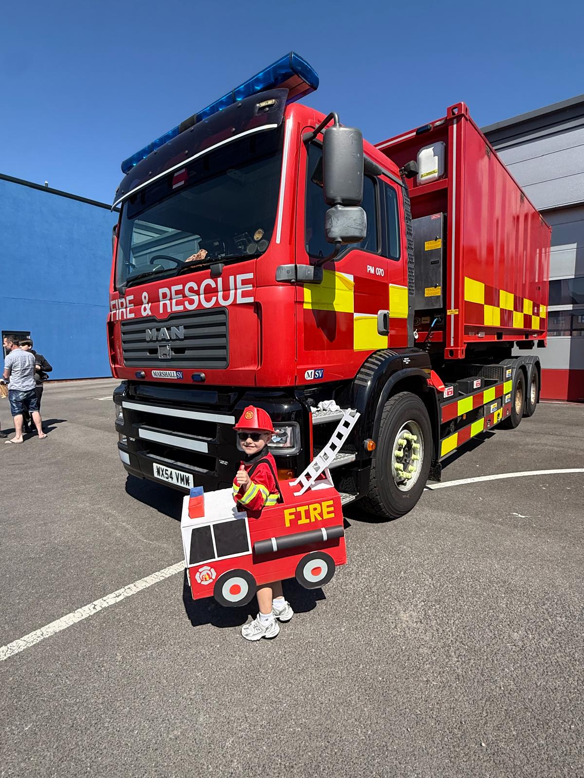 Young boy dressed as a fire truck standing in front of fire appliance in yard